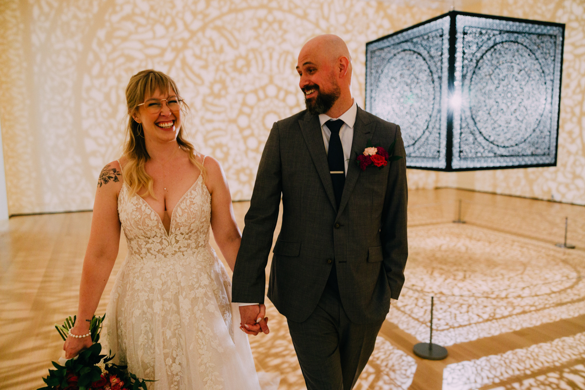 A joyous bride and groom hold hands, smiling at each other amidst intricate shadow patterns created by artwork in a spacious venue.