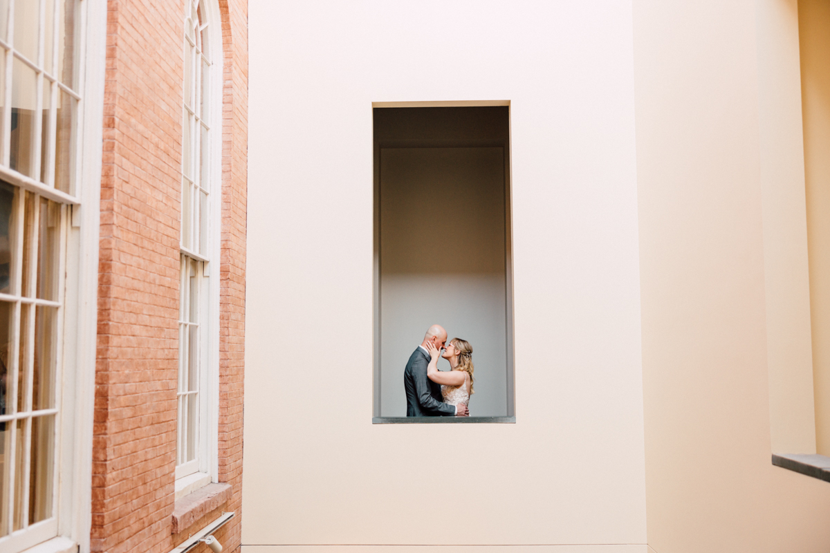 A couple shares a tender moment in a minimalist architectural space, framed by soft pastel walls.