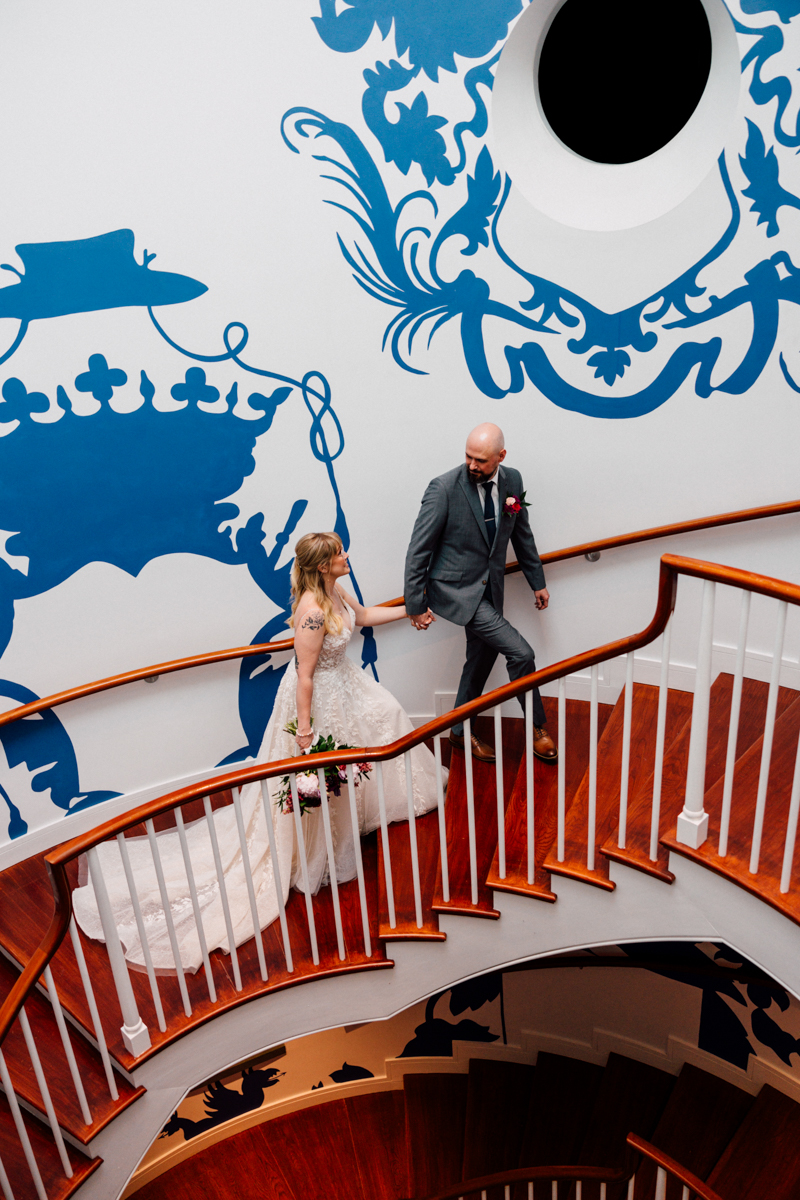 A couple ascending a beautifully designed staircase, adorned with blue wall art, celebrating their wedding.