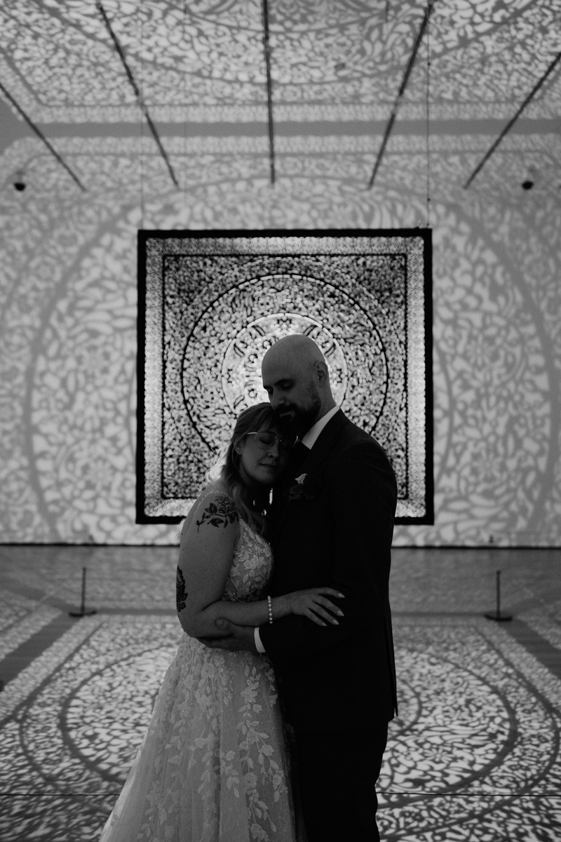 A couple embraces in a dimly lit art space, surrounded by intricate patterned shadows, capturing a moment of love and serenity.