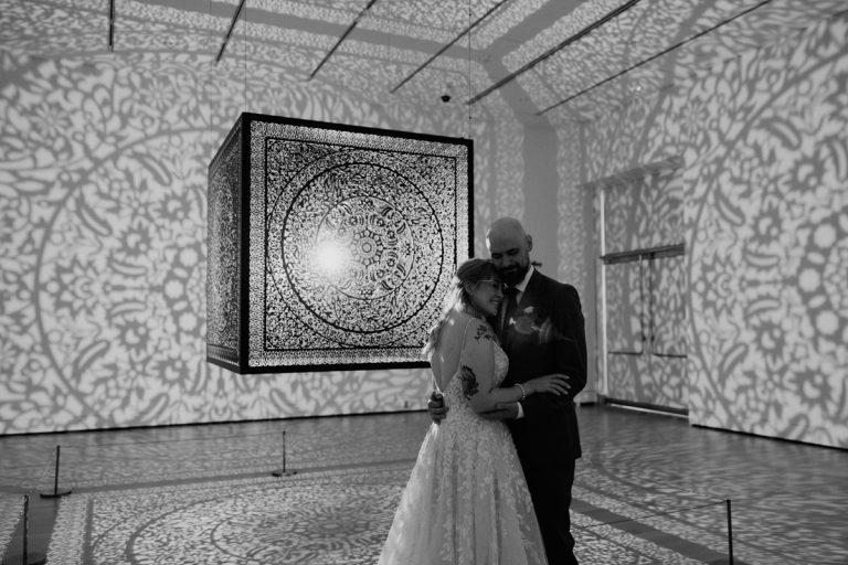 A couple embraces in an art installation space, surrounded by intricate shadows cast by a suspended patterned cube.