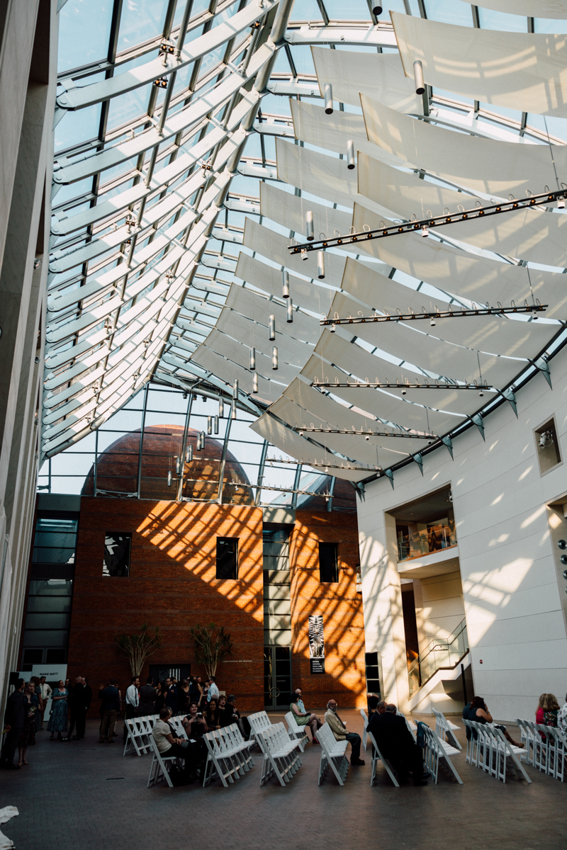 An interior view of a modern building featuring a glass roof, fabric awnings, and natural sunlight casting patterns on the walls and floor, with people seated in folding chairs.