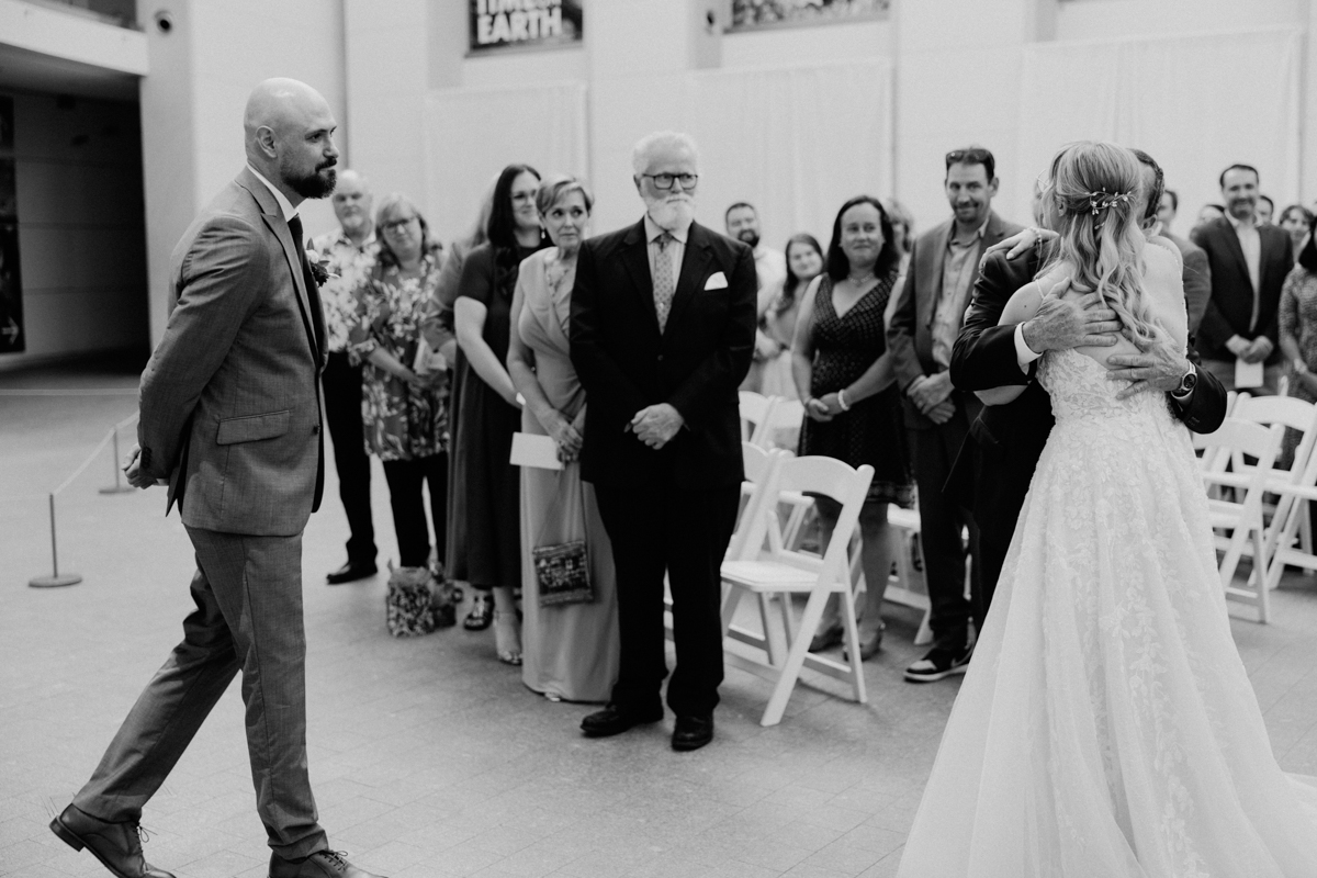 A groom stands with his back to the camera as a bride embraces her father, surrounded by attentive guests in a wedding venue.