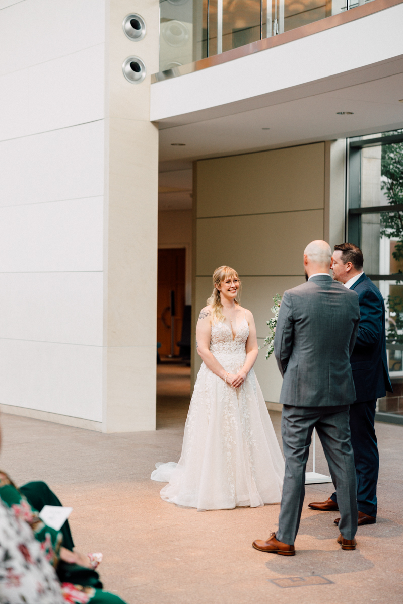 A bride smiling during her wedding ceremony, standing before two men, in a spacious and elegantly designed indoor setting.