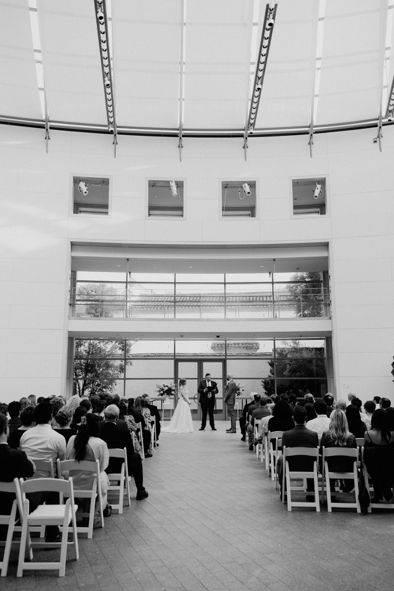 A couple exchanges vows during a wedding ceremony in a modern indoor venue, surrounded by seated guests.