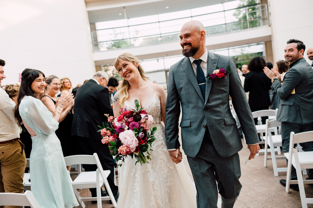 A newlywed couple smiles and holds hands while walking down the aisle at their Peabody Essex Museum Wedding, surrounded by applauding guests. The bride holds a vibrant bouquet, and both look joyful in a bright indoor venue.