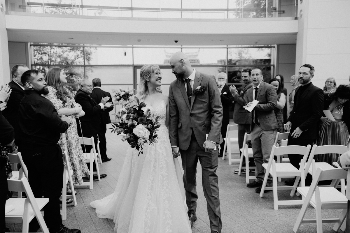 A bride and groom smile at each other while holding hands and walking down the aisle at their Peabody Essex Museum Wedding. Guests stand, clap, and capture the moment in a bright, modern venue filled with joy.