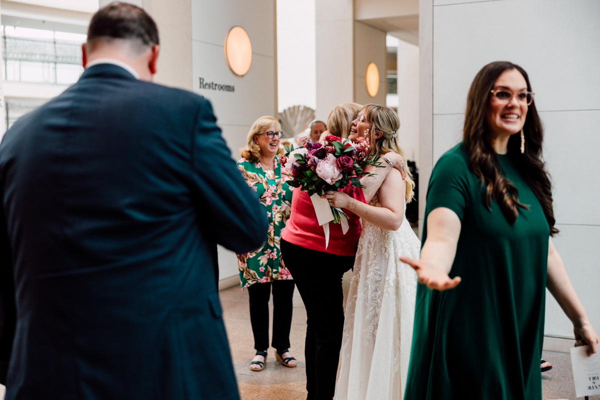At a Peabody Essex Museum wedding, a bride in a white dress hugs a woman in a pink top while holding her bouquet. Three others stand nearby, including a woman in green reaching out and a man in a suit. Restrooms sign is visible in the background.