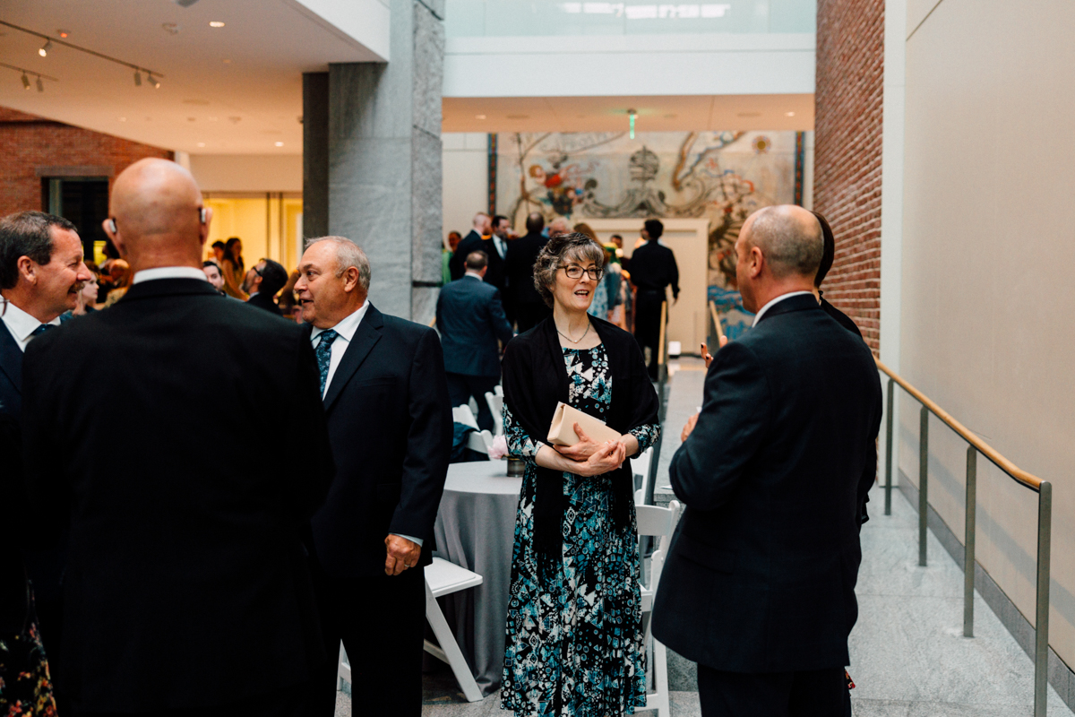 A group of people in formal attire stand and converse in a bright atrium with marble floors and brick walls at a Peabody Essex Museum Wedding. A woman in a floral dress holds a book, smiling, while others chat near round tables.