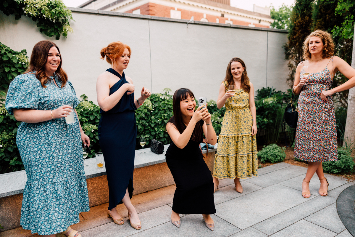 Five women in colorful dresses stand outdoors, smiling and laughing at a Peabody Essex Museum Wedding. One woman crouches to take a photo with her phone, while the others pose and react playfully in a garden-like setting.