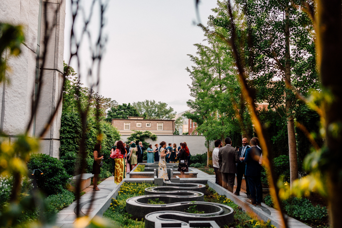 A group of people dressed in formal attire gather and mingle in a lush garden courtyard at a Peabody Essex Museum Wedding, with geometric water features surrounded by green trees and shrubs on a clear day.