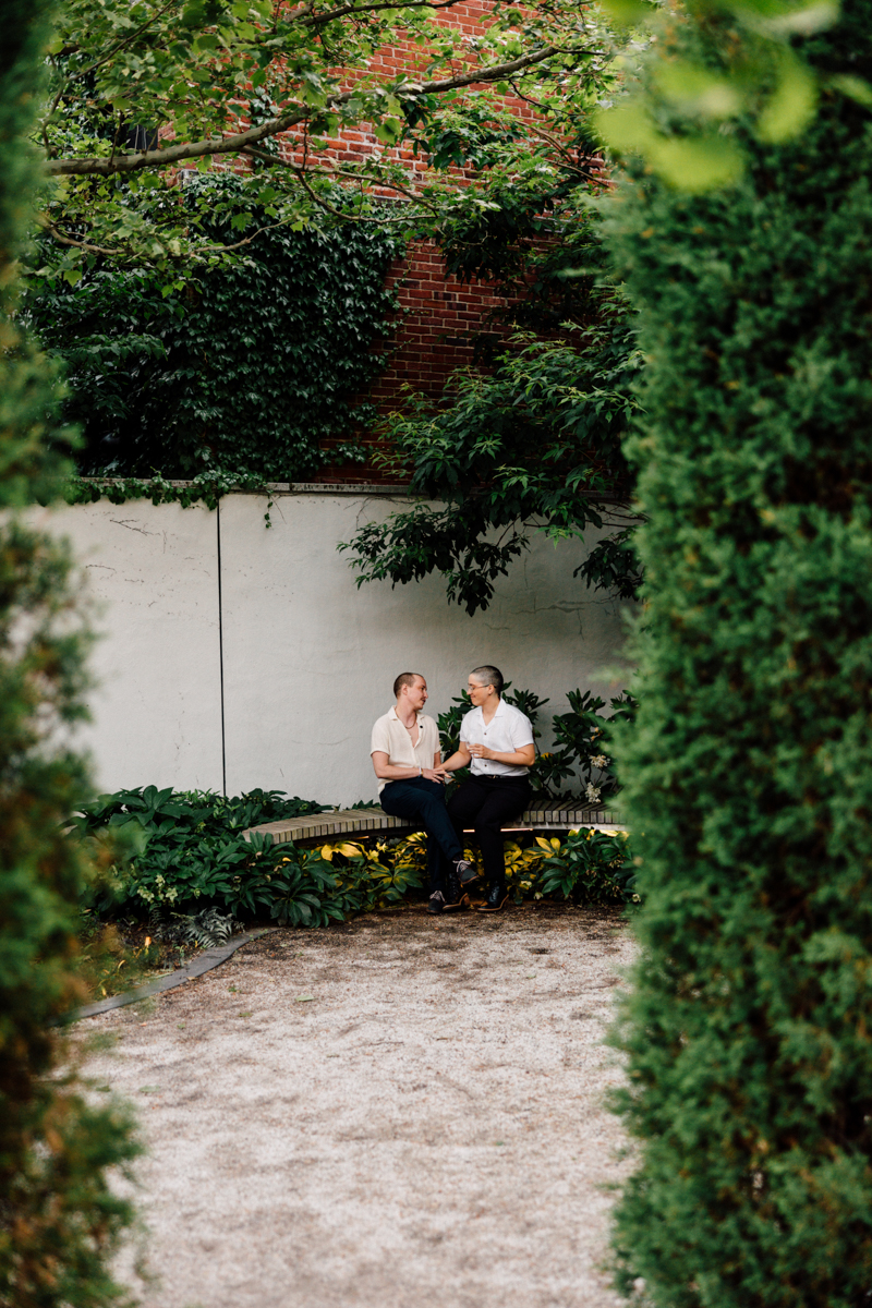 Two people sit together on a bench in a lush, green garden at the Peabody Essex Museum Wedding, facing each other and smiling while holding hands. Tall trees and dense foliage frame the scene, creating a peaceful, intimate atmosphere.