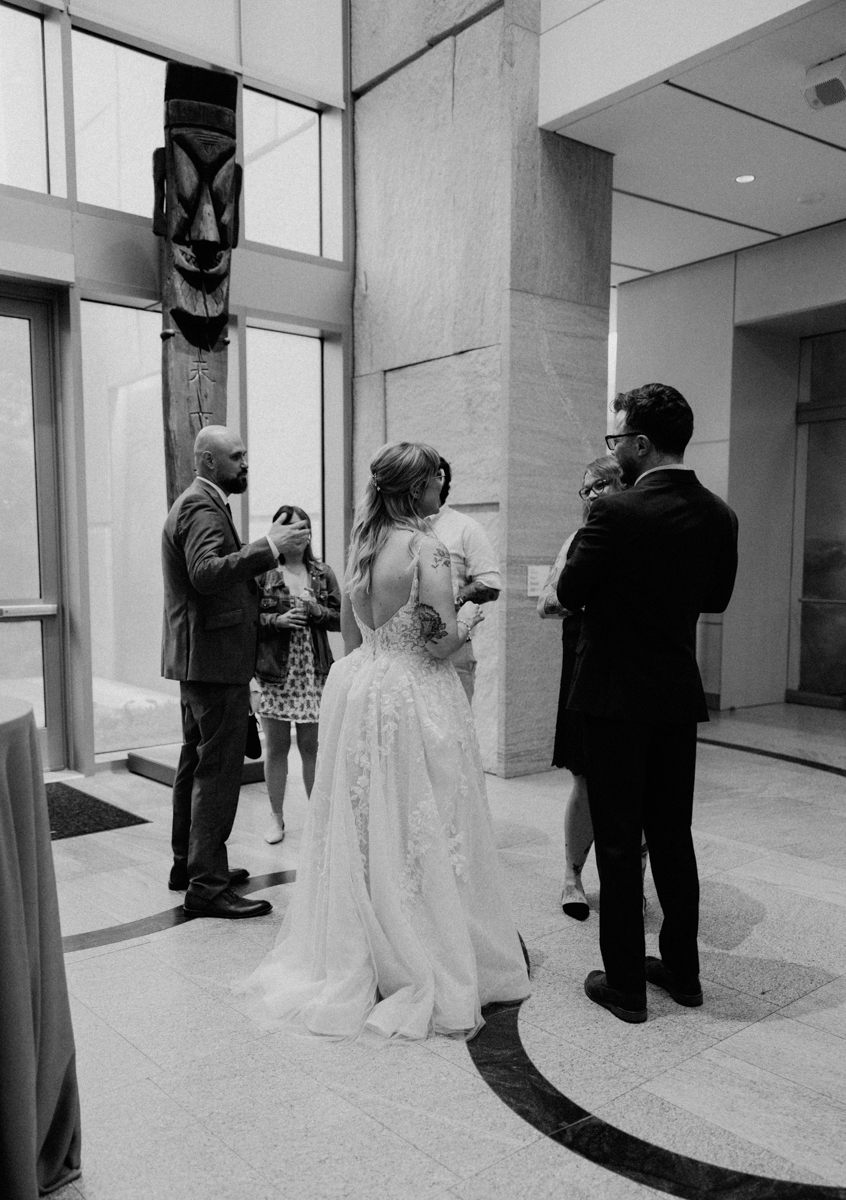 A bride in a lace dress stands with four guests in formal attire, conversing in a modern, spacious indoor setting with tall windows and stone walls at a Peabody Essex Museum Wedding.