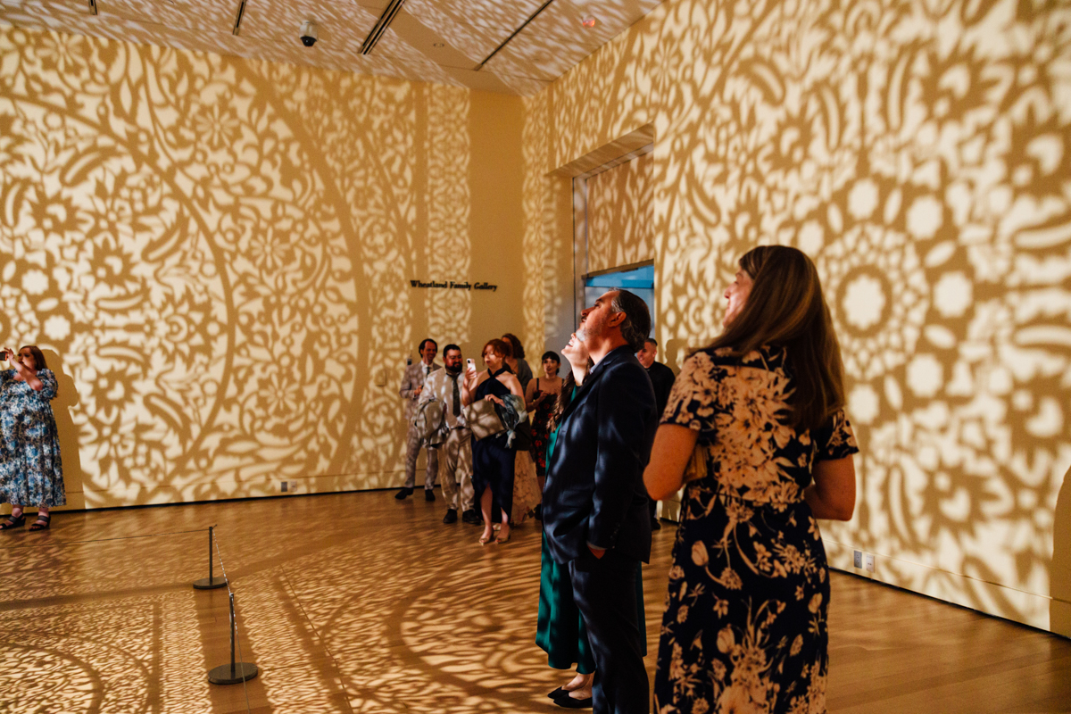 People stand in a gallery at the Peabody Essex Museum Wedding, intricate lace-like shadows cast across the walls and ceiling by an elaborate light installation. Some guests gaze upward, admiring the display, while others chat or take photos.
