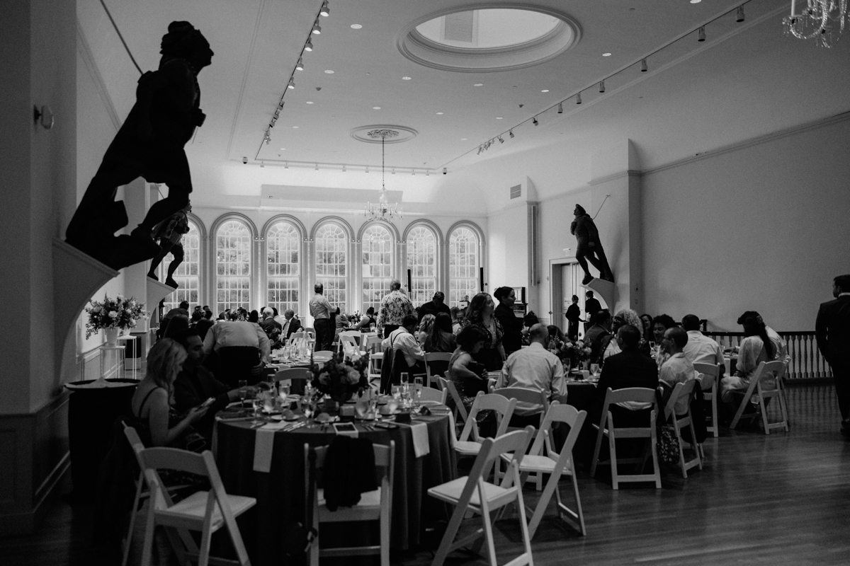 A black-and-white photo of a formal Peabody Essex Museum Wedding in a large, elegant room with arched windows and statues. Guests dine and converse at round tables beneath high ceilings and stylish track lighting.