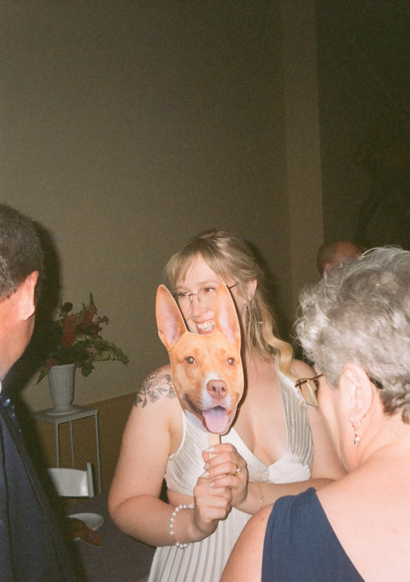 A woman in a white dress holds a cutout of a dog’s face over her own, smiling as she stands among other guests at a Peabody Essex Museum wedding. Flowers in a vase are visible in the background.