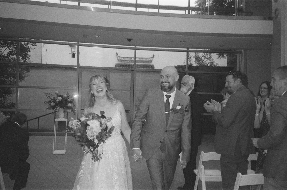 A bride and groom walk hand in hand, beaming with joy as guests applaud their marriage in a beautifully lit venue.