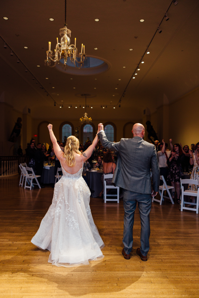 A bride and groom hold hands, raising them in celebration as they step into a warmly lit venue filled with guests in an elegant setting.