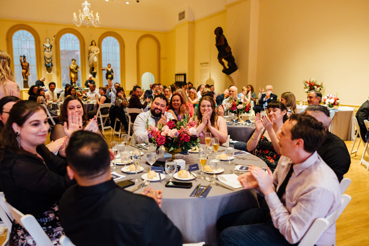 Guests enjoying a lively moment, clapping and smiling during a formal dinner event, surrounded by elegant table settings and floral arrangements.