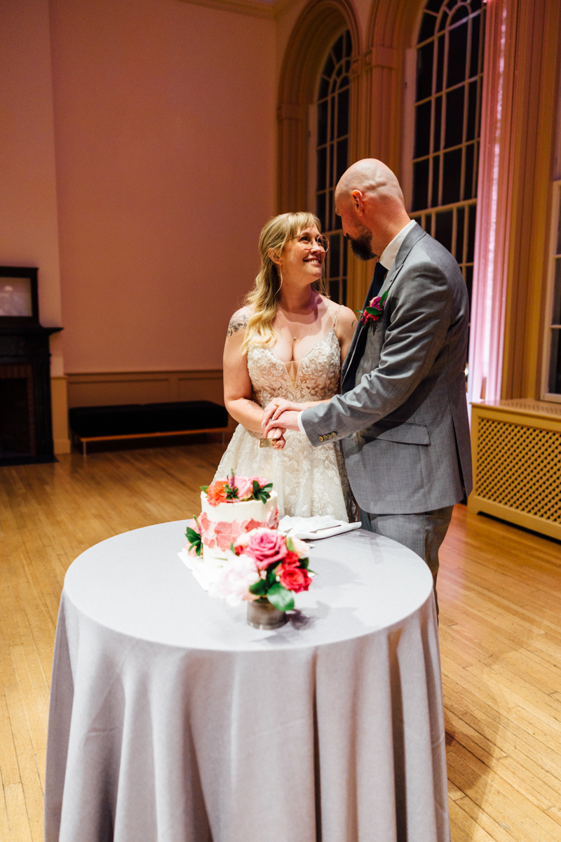 A joyful bride and groom share a loving gaze during their wedding ceremony, standing by a decorated table with a small wedding cake and flowers.