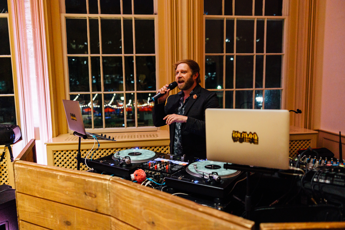 A DJ energetically singing into a microphone while spinning records at a night event, with a view of lights outside through large windows.