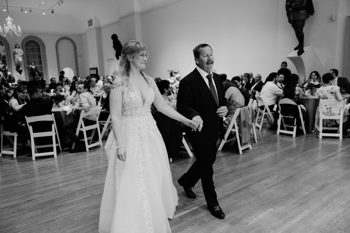 A bride and her father walking hand in hand into a wedding reception, with guests seated at tables in the background.