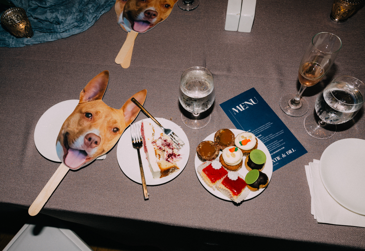 A top-down view of a table featuring dog face masks, various desserts on plates, a menu, and drinks, all set against a textured tablecloth.