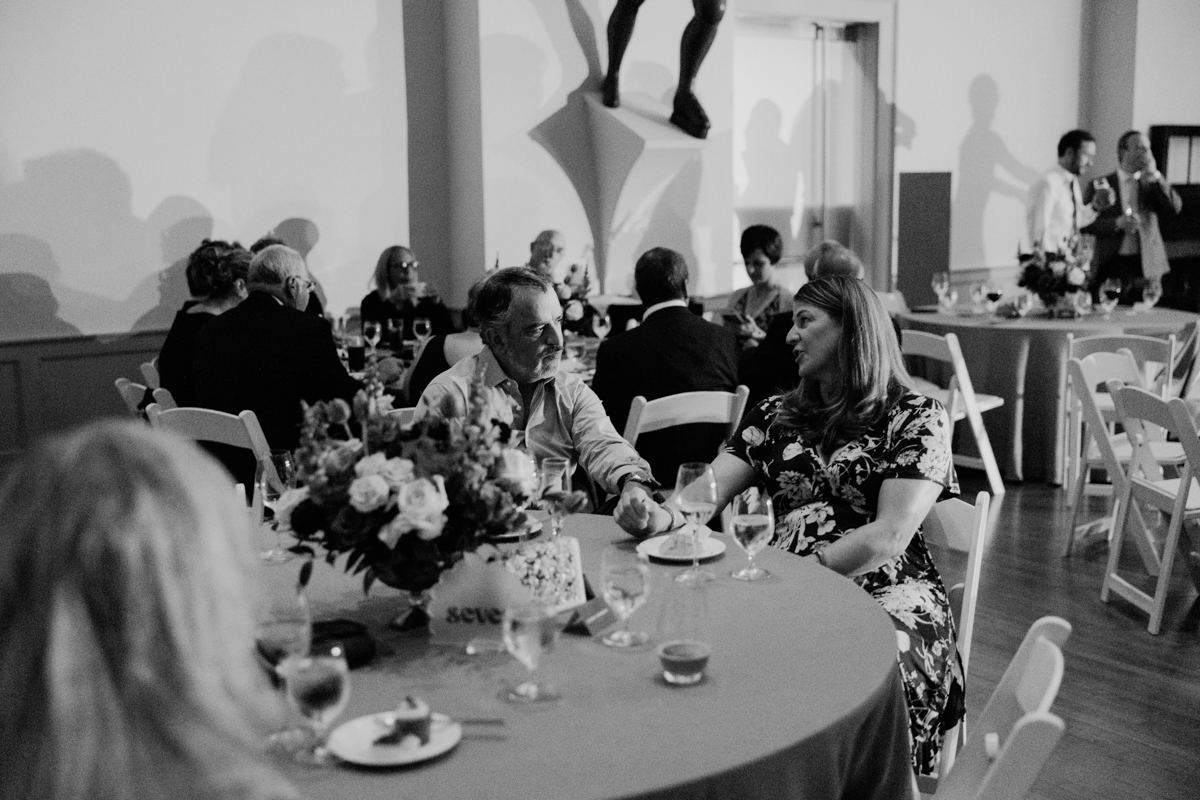 A black and white photo of guests enjoying a seated event, sharing conversations around elegantly set tables with floral centerpieces.