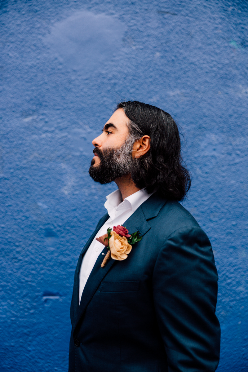 A man with dark hair and a beard, dressed in a dark suit and white shirt with a boutonnière, stands in profile against a textured blue wall—an elegant portrait inspired by Cambridge City Hall Elopement Photography.
