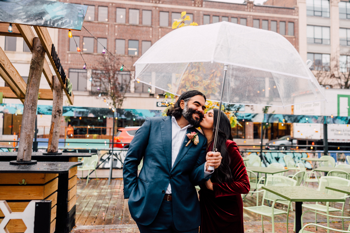 A smiling couple stands close under a clear umbrella on a rainy day. The woman in a red velvet dress kisses his cheek as he leans in, dressed in blue—capturing the romance of Cambridge City Hall Elopement Photography with café tables and string lights behind them.
