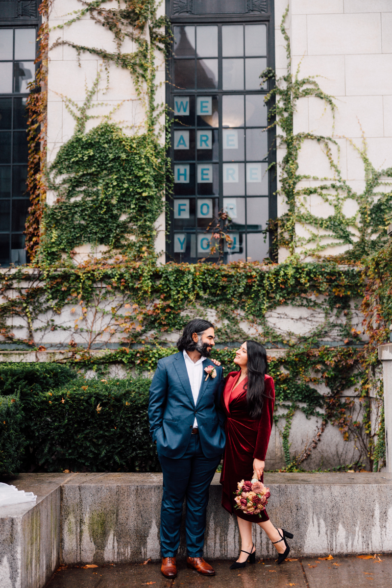 A couple stands in front of a vine-covered building, smiling at each other. The man wears a blue suit; the woman is in a red dress holding a bouquet—a charming moment captured by Cambridge City Hall Wedding Photography.