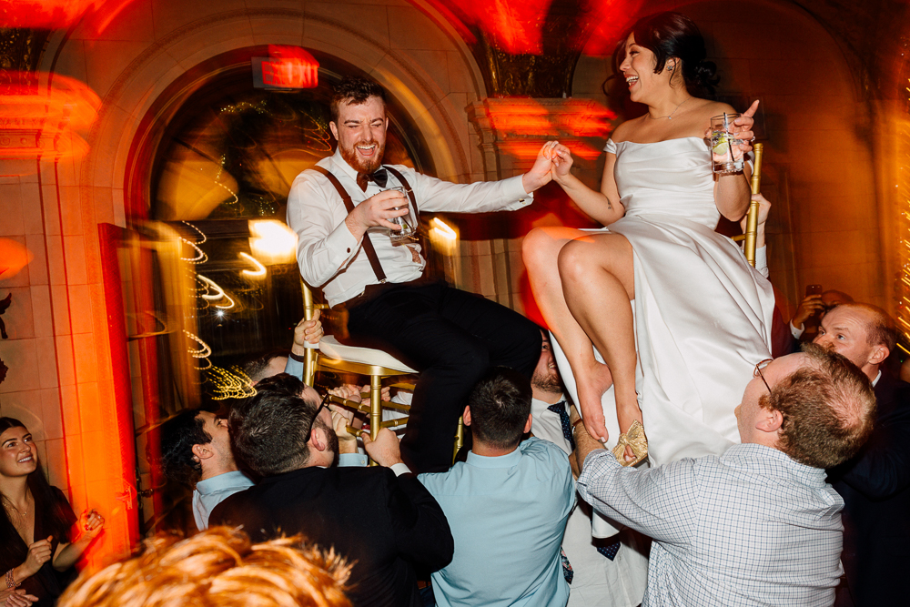 A joyful bride and groom are lifted on chairs by guests during a lively Aldrich Mansion Wedding Photography celebration indoors, surrounded by smiling friends under warm, glowing lights.