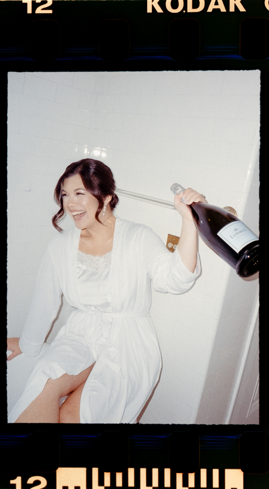 A woman in a white robe sits in a bathtub, smiling and holding up a large bottle of champagne. The image, reminiscent of Aldrich Mansion Wedding Photography, features a film border and exudes a light, candid feel.