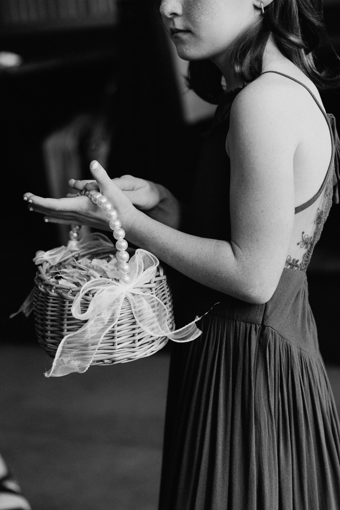 A young girl in a sleeveless dress holds a wicker basket decorated with a ribbon and pearls, while scattering flower petals with her other hand. The image is in black and white.