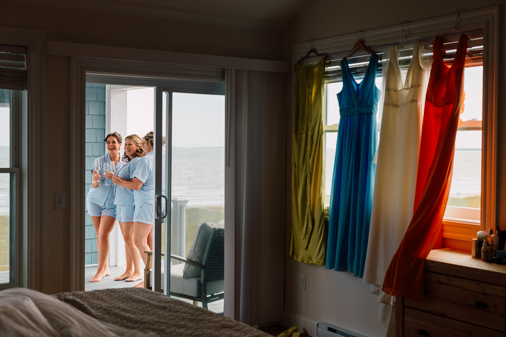 Three women in matching blue pajamas stand on a balcony by the ocean, laughing and holding drinks. Inside, four colorful dresses—yellow, blue, white, and orange—hang in front of a window.