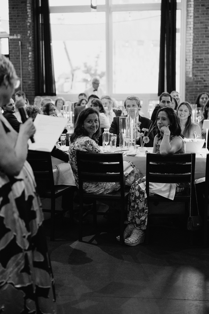 A group of people sit at round tables, attentively listening to a person speaking and holding a paper at the front. The setting is indoors with large windows letting in natural light. The mood appears formal and engaged.