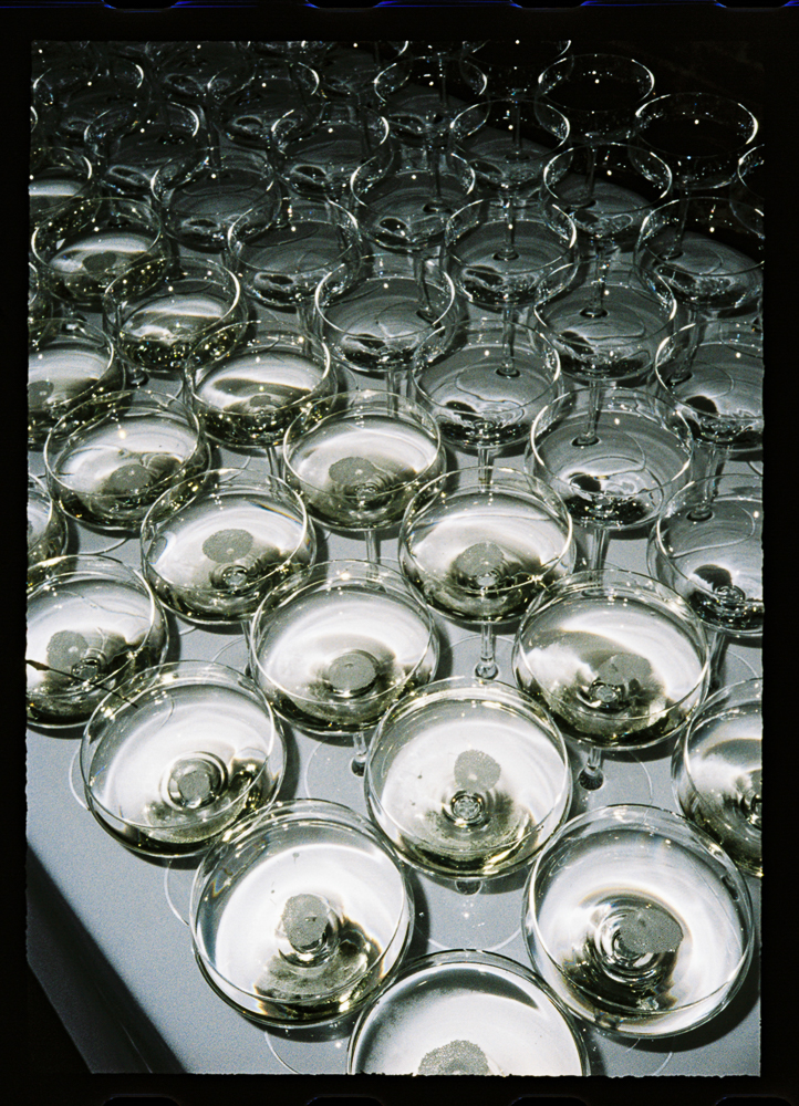 Rows of empty champagne glasses arranged closely together on a white surface, viewed from above, creating a repeating pattern with reflections and highlights in the glass.