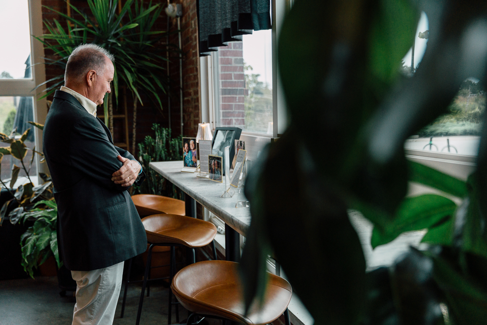 A man in a suit stands by a bright window, looking at framed photos on a counter in a room filled with green plants and wooden chairs.