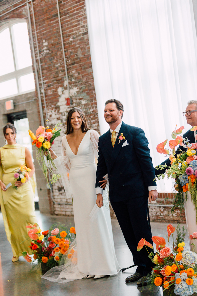 A bride in a white gown and a groom in a dark suit stand smiling at an indoor wedding ceremony, surrounded by colorful floral arrangements. A bridesmaid in a yellow dress stands nearby. Natural light streams through large windows.