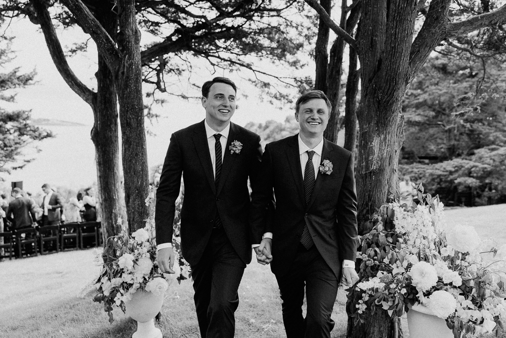 Two men in suits, smiling and holding hands, walk between floral arrangements at an outdoor wedding ceremony, surrounded by trees and greenery.