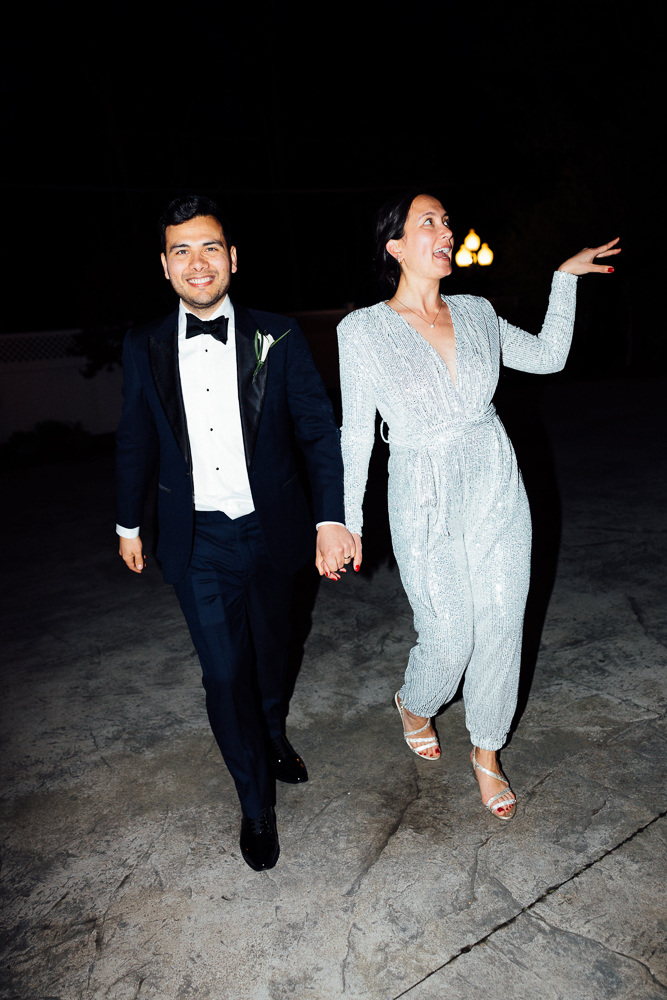 A smiling couple holds hands and walks outside at night. The man wears a black tuxedo with a bow tie, and the woman wears a sparkly silver jumpsuit with high heels, gesturing joyfully with her free hand.