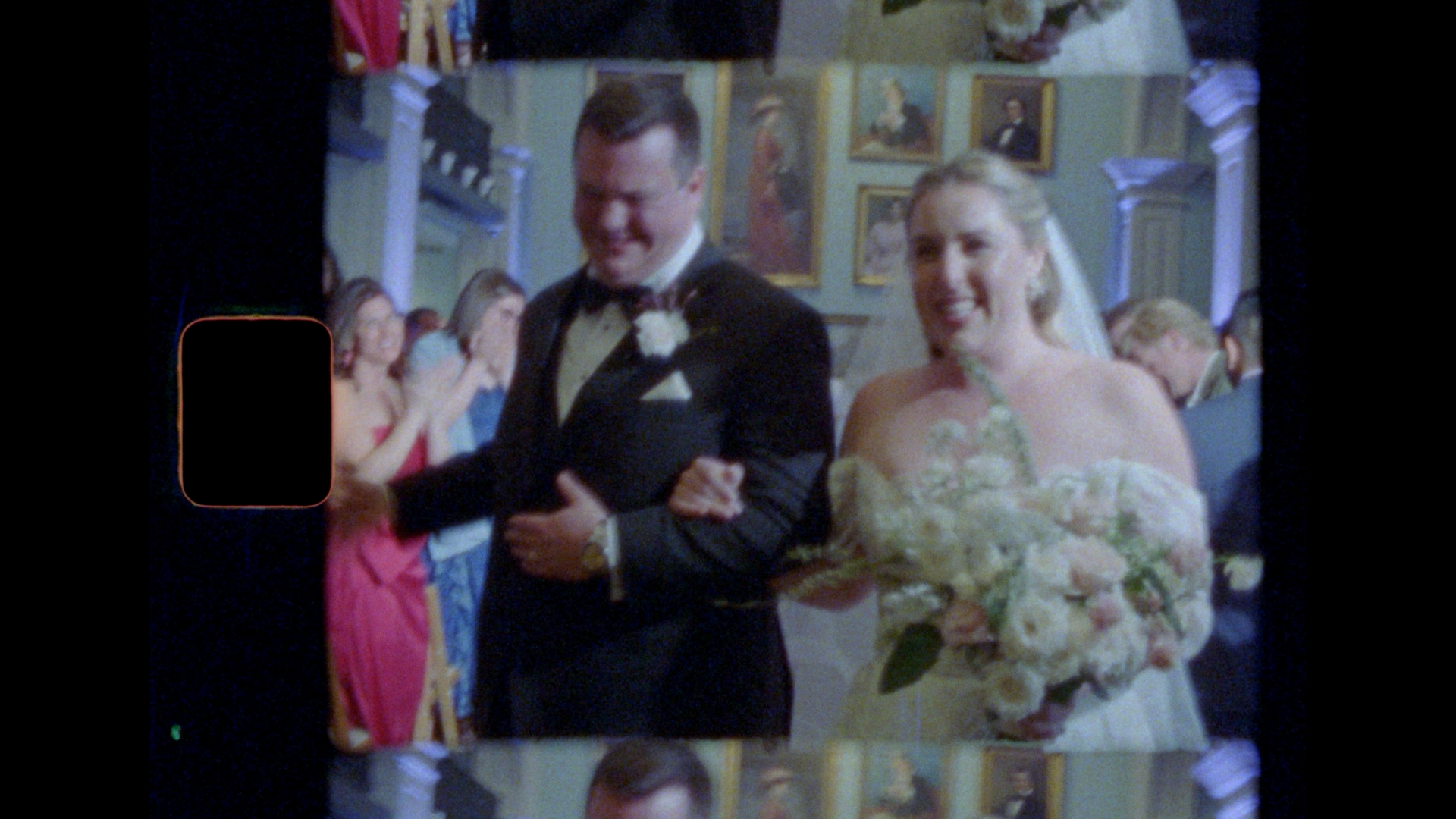 A bride and groom, smiling and arm in arm, walk down the aisle surrounded by guests. The bride holds a large bouquet of flowers, and the groom wears a black tuxedo. The image has a vintage, film-like quality. It was taken on Super 8 Film