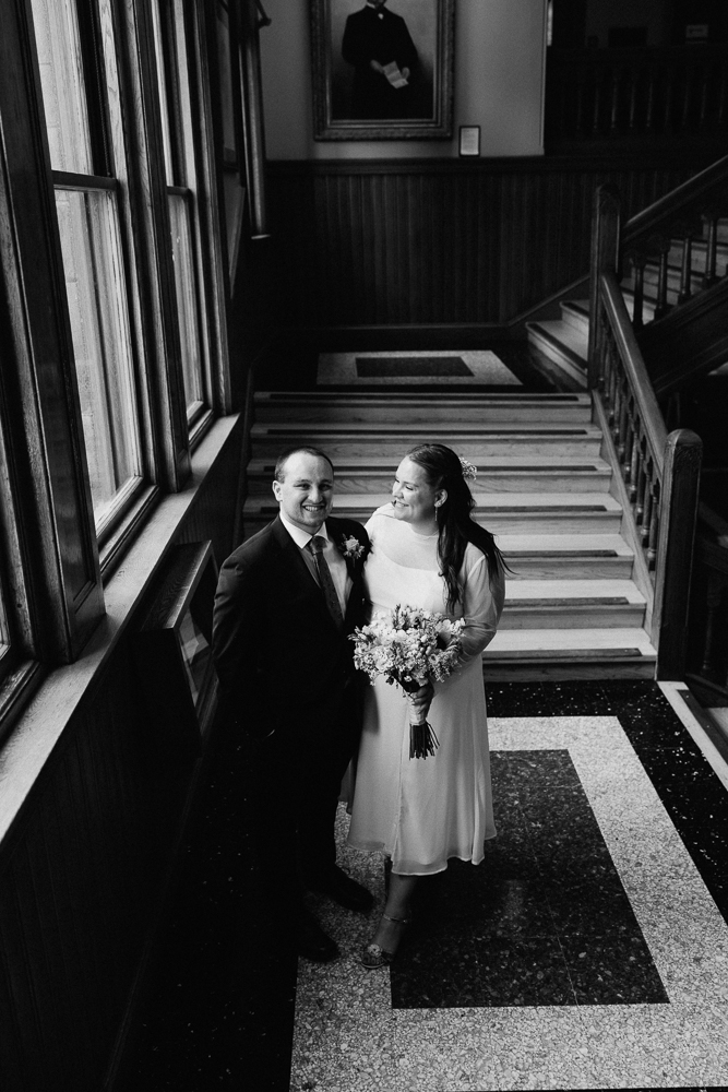 A black-and-white photo of a smiling couple in wedding attire standing by a staircase in a classic building. The bride holds a bouquet of flowers, and sunlight streams through large windows beside them.
