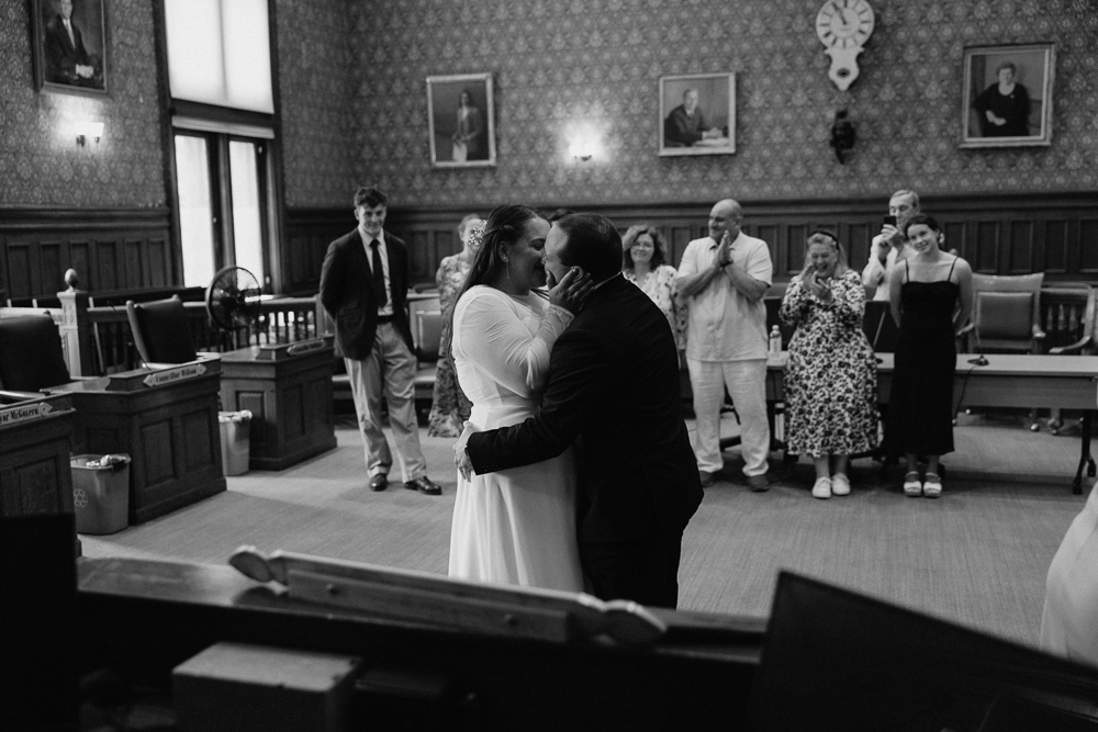 A bride and groom kiss in a courtroom, surrounded by clapping and smiling guests. The scene is joyful, with people standing and celebrating the couple’s wedding ceremony. The image is in black and white.