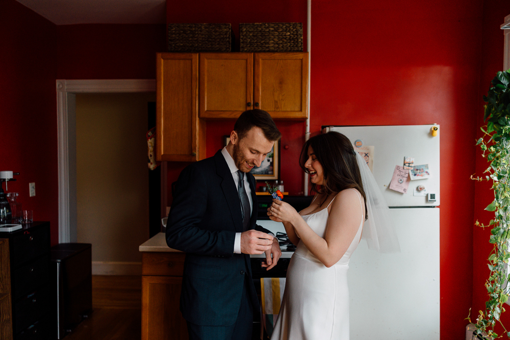 A bride in a white dress and veil laughs while handing a small object to a groom in a suit. They stand together in a kitchen with red walls and wooden cabinets, sharing a joyful moment.