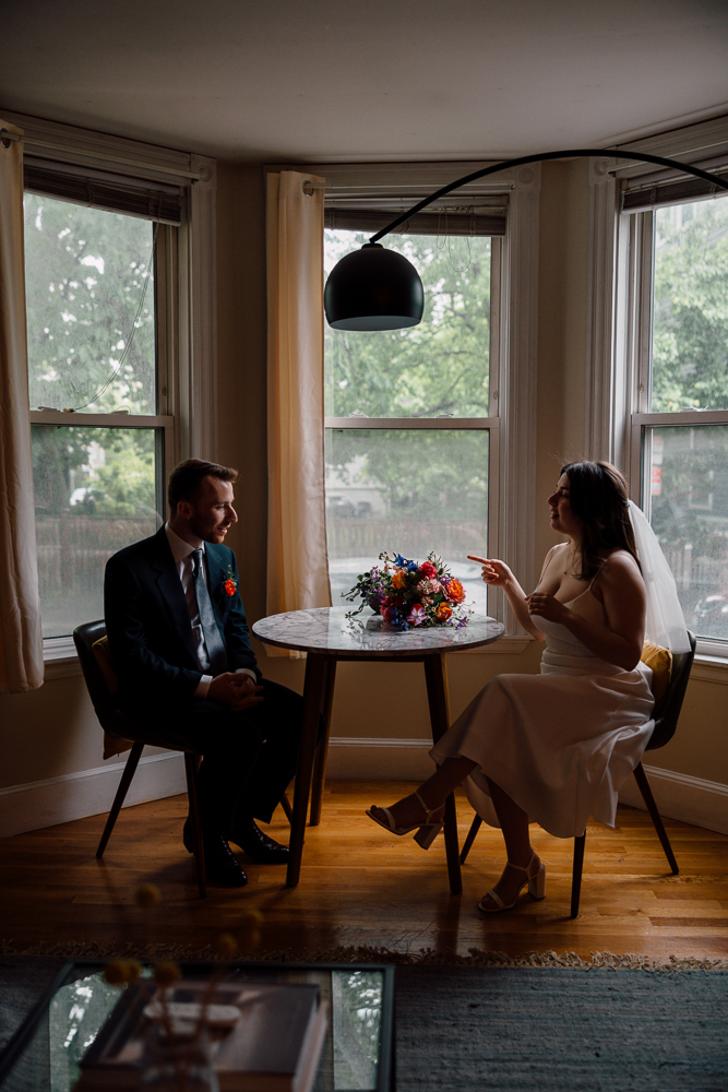 A bride and groom sit across from each other at a small round table by large windows in a softly lit room. The bride gestures with one hand, and a bouquet of colorful flowers lies on the table between them.