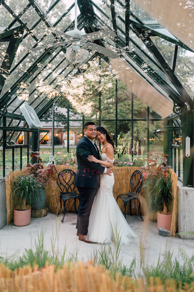 A bride and groom embrace and smile inside a glass greenhouse decorated with potted plants and string lights. Natural light streams in around them, creating a warm, romantic atmosphere.