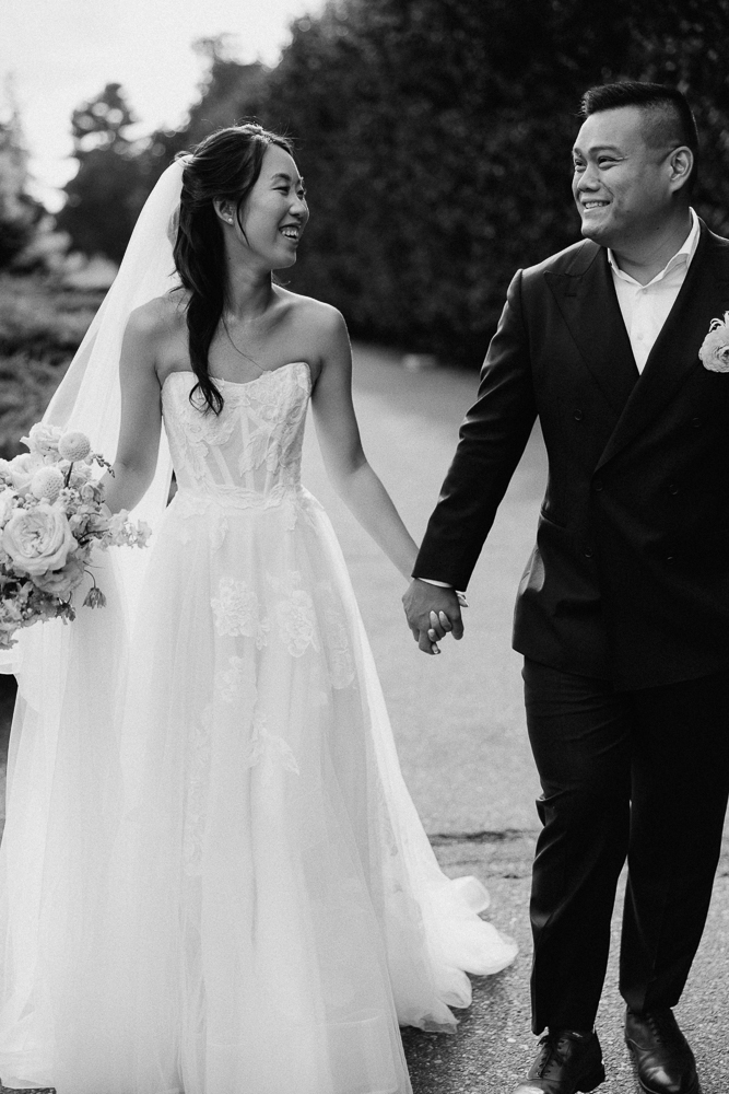 A smiling bride in a strapless lace gown and veil holds a bouquet and the hand of a groom in a dark suit as they walk outdoors, looking happily at each other.