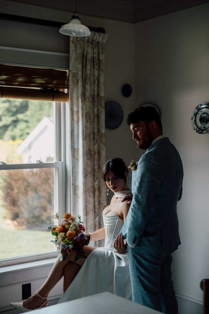 A bride in a white dress sits by a window holding a colorful bouquet, while a groom in a light blue suit stands beside her. Soft daylight streams in, creating a warm, intimate atmosphere in the room.