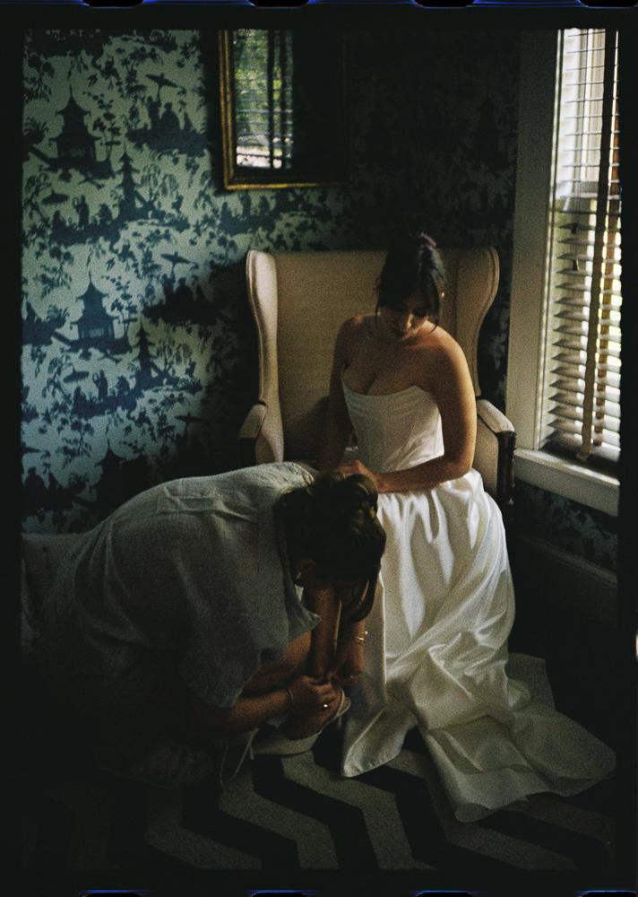 A bride in a strapless white gown sits on a chair in a softly lit room while another person kneels, helping her with her shoes. The room has patterned wallpaper, a window with blinds, and a chevron rug.
